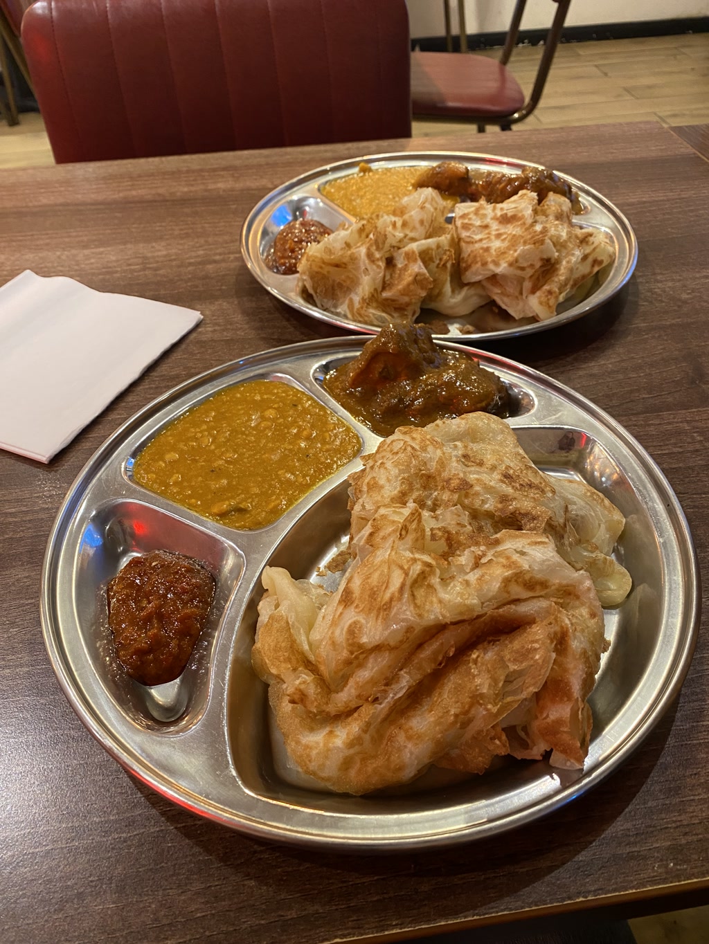 A meal consisting of flaky, layered flatbread served on a metal tray alongside various types of curry and condiments. The flatbread appears golden brown and crispy, likely a type of Roti or Paratha. The curries are served in compartments on a second metal tray and range in color from yellow to dark brown, suggestive of different spices and ingredients. Additionally, there are small portions of condiments that might be spicy sambal or chutney, complimenting the main dish. The setting looks like a simple dining area with a wooden table and a red vinyl upholstered bench in the background.