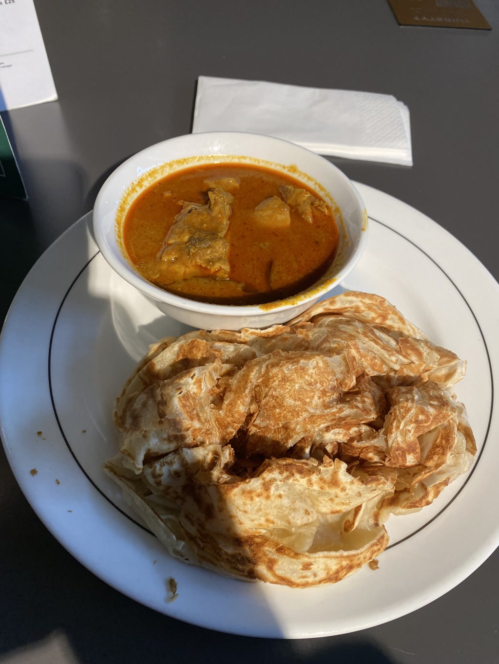 A bowl of curry with large pieces of meat is accompanied by a portion of flaky, layered flatbread on a white plate. The curry appears rich and vibrant with a reddish-orange color, indicating a blend of spices. The flatbread, known as roti canai or roti paratha, is golden brown with crisp, uneven surfaces, suggesting it is freshly pan-fried to achieve a combination of crunchy and soft textures. Both items are served on a dark tabletop, with a white napkin and a corner of a document or menu seen in the backdrop.