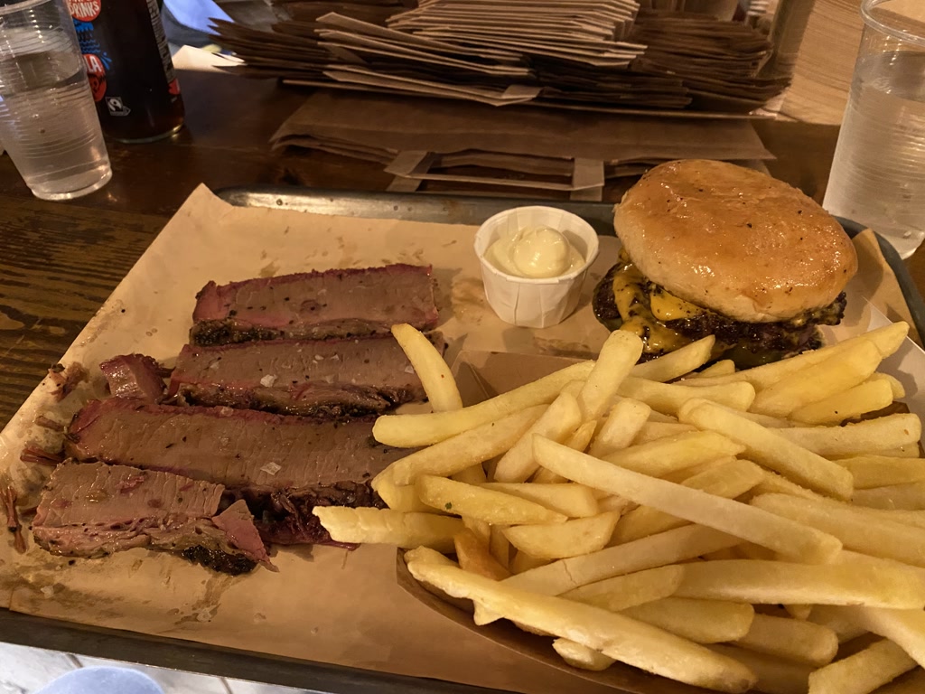 A meal consisting of sliced smoked brisket, a side of french fries, and a cheeseburger with pickles. The burger is on a sesame seed bun with a side of mayonnaise. The brisket is arranged neatly on parchment paper and shows a pink smoke ring, indicating it has been smoked. The fries are golden in color and appear crispy. There is a can of sparkling water in the background as well as stacked brown paper bags or menus, suggesting this meal is being enjoyed in a casual dining restaurant.