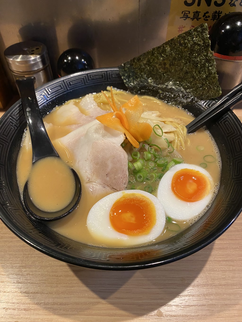 A bowl of ramen with a creamy, possibly tonkotsu-based broth. The bowl contains sliced green onions, two halves of a soft-boiled egg with a semi-solid yolk, slices of tender-looking pork, a single piece of nori (seaweed), and possibly bamboo shoots or sliced fish cake arranged artistically. The ramen noodles are not clearly visible but are likely submerged in the broth. A black spoon and chopsticks rest on top of the bowl, and the tableware has a traditional Asian design. A condiment shaker can be seen in the background, and there's a partial view of a yellow poster or sign with Japanese characters that are not fully visible and therefore the text cannot be fully extracted.