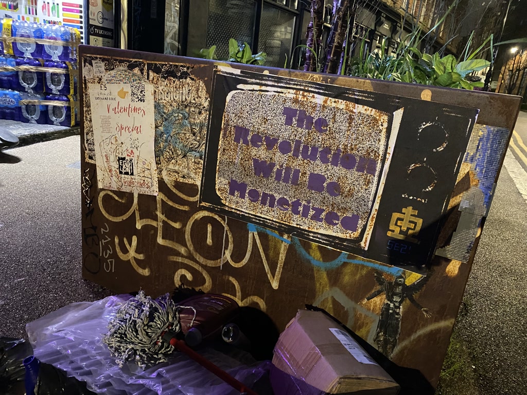 A large, worn street cabinet covered in various stickers, graffiti, and posters stands on a sidewalk at night. One prominent section is stenciled with glittery text on a rusted background. A small stencil below the text illustrates a figure holding an object aloft, resembling a flag or a torch. The cabinet sits beside potted plants that appear to be part of an outdoor display near a retail setup, with stacks of bottled water visible in the background. Refuse, including a shredded piece of cloth and a purple, taped parcel, is scattered at the base of the cabinet.