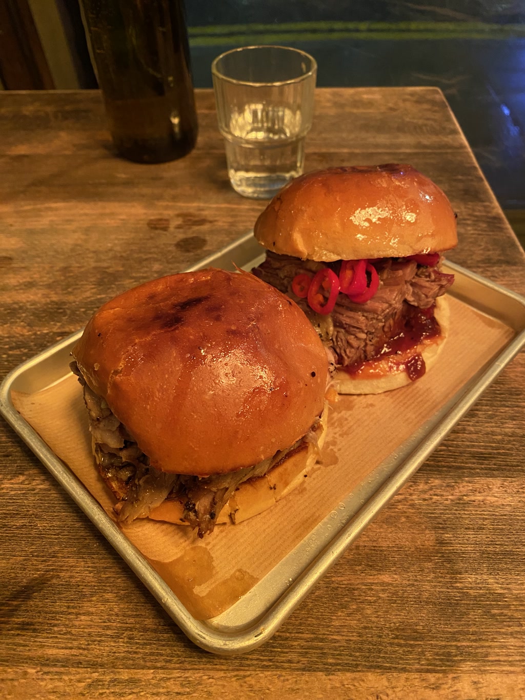 The photo shows two sizeable burgers served on a metal tray with a piece of parchment paper underneath. The buns appear shiny and toasted, indicating they might be brioche buns. The burger in the foreground has a generous amount of caramelized onions on top of a well-cooked piece of meat, which could likely be pulled pork or beef, based on the texture. The other burger, slightly behind the first, features vibrant pickled red onions atop the meat, adding a pop of color and likely a tangy flavor. Side items are not visible in the view. In the background, a bottle and a half-full glass of a clear liquid, possibly water, are present on the rustic wooden table.