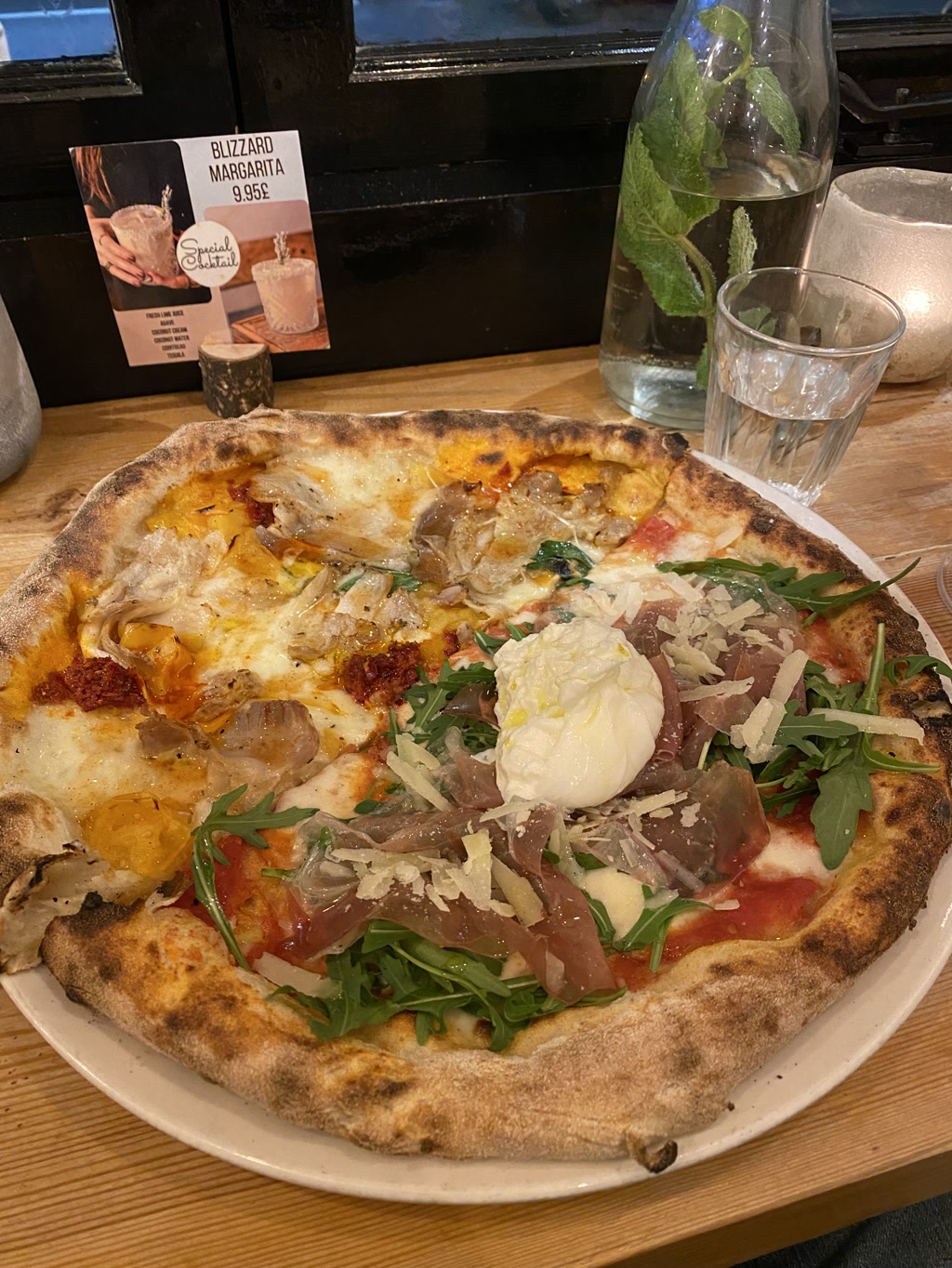 A freshly baked pizza is presented on a round white plate, exhibiting a golden-brown crust with char marks. The pizza is topped with a variety of colorful ingredients, including melted cheese spread unevenly across the surface, bright red tomato sauce visible in areas, vibrant green arugula, a few slices of what appears to be prosciutto, and dollops of yellow and red condiments that could be different types of flavored oils or sauces. Additionally, there is a visibly soft, poached egg on top with a-runny yolk. Set beside the pizza on the wood-finished table, there is a clear glass of water, a bottle of water with mint leaves inside, and a candle holder with a lit candle giving off a soft glow. A promotional card for a 'Blizzard Margarita' is propped up against the window, suggesting the venue might be a restaurant or cafe with a focus on Italian cuisine or a selection of specialty drinks.