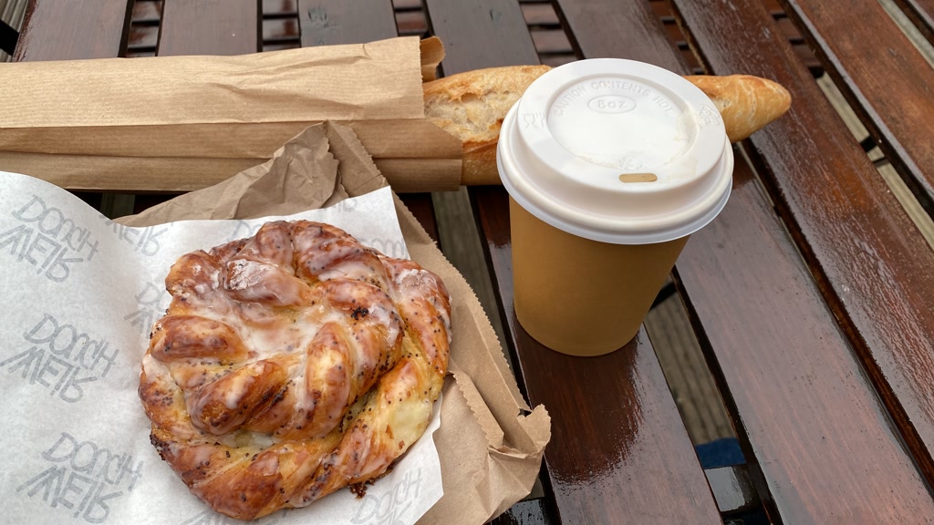 A breakfast scene with a cup of coffee in a take-out paper cup with a plastic lid on the right. Next to the coffee, there's a pastry with icing on parchment paper with a branded logo. On the left side of the photo, a paper bag contains a partially visible baguette. The items are placed on a wooden slatted table that appears to be outdoors, as indicated by the wet surface suggesting recent rain or outdoor conditions.