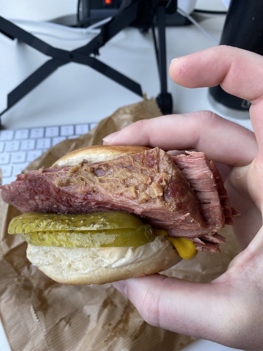 A hand is holding a sandwich consisting of a large slice of brisket and a single pickle on a white bun. The brisket appears well-cooked with a juicy texture, and the pickle is bright green indicating a pickled cucumber. The setting is an office environment with a computer keyboard, a paper bag, and part of a tripod stand visible in the background.