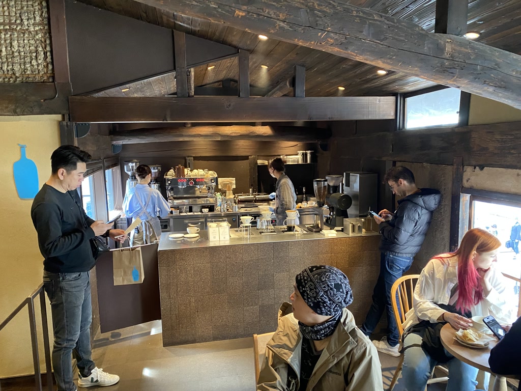 Inside a cozy and rustic café with exposed wooden beams, a group of patrons engage in various activities. On the left, a man stands near the entrance while looking at his phone, holding a shopping bag. In the center, two individuals are standing at the coffee bar; one appears to be a barista preparing a drink, while the other is interacting with a smartphone. To the right, seated guests include a person in a trench coat facing away from the camera and a woman with red hair focused on her phone. The interior has a warm ambiance with natural light streaming in from the windows.