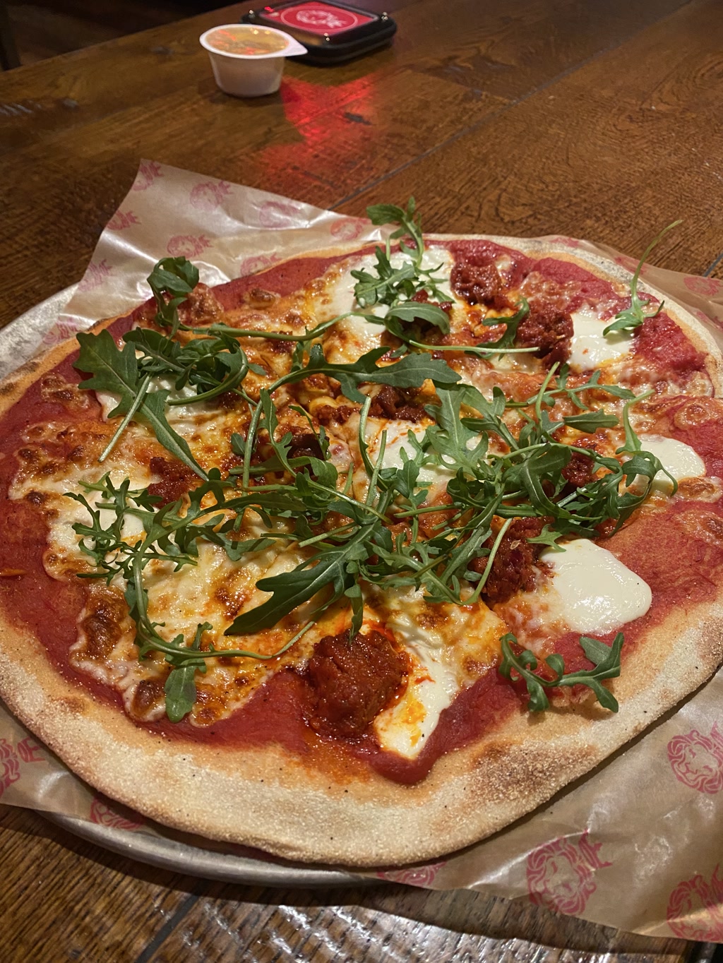 A whole pizza with a slightly charred crust is presented on a table, topped with vibrant green arugula leaves scattered over melted cheese with golden-brown spots. There are pieces of what appears to be spicy salami or pepperoni and dabs of white, possibly a soft cheese like mozzarella or ricotta. A container of dipping sauce is on the side, and a circular electronic device, maybe a restaurant pager, is in the background on the wooden table.